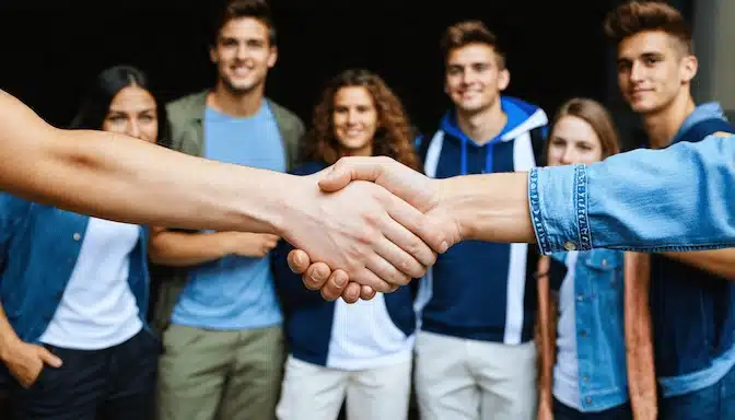 Deux personnes se serrent la main devant un groupe de jeunes souriants, symbolisant l’accueil et la convivialité lors de la journée portes ouvertes de la formation NTC.